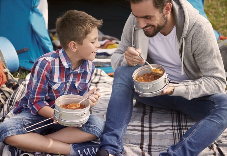 Pappa och son äter varm soppa ur matburkar av stål vid ett tält